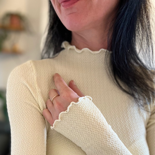 Woman wearing a beige textured sweater with a blurred indoor background with a 9ct gold ring on her hand.